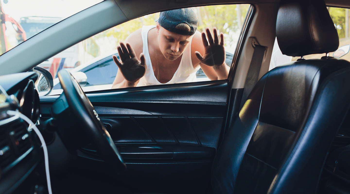Man in a white tank top and backward cap pressing his hands against a car window from outside.