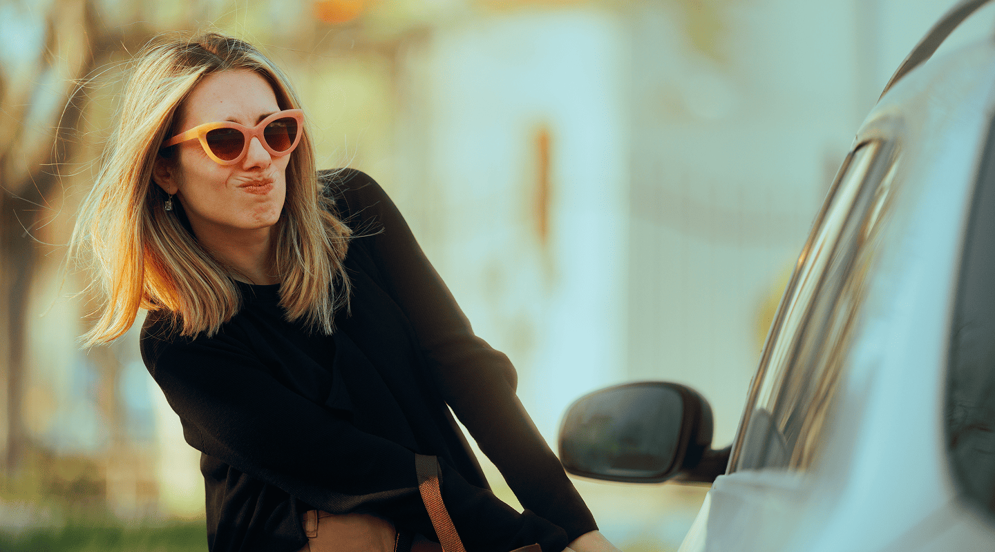Young woman with blonde hair and orange sunglasses making a playful face while holding the door handle of a white car.